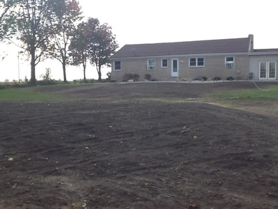 Single-story beige house with a brown roof, front yard mostly bare soil and a few trees on the left. - arwwm - arwwm