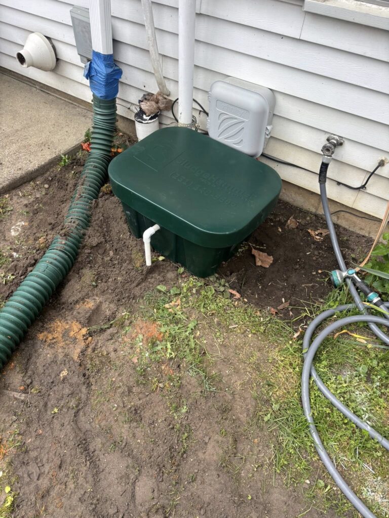 492768382_1320299279632911_2641541054826008890_n - ARWWM - ARWWM Green plastic rain barrel with lid sits near the house siding, with a downspout and blue tape, plus a long green drainage hose on bare soil and a garden hose nearby. - arwwm - arwwm
