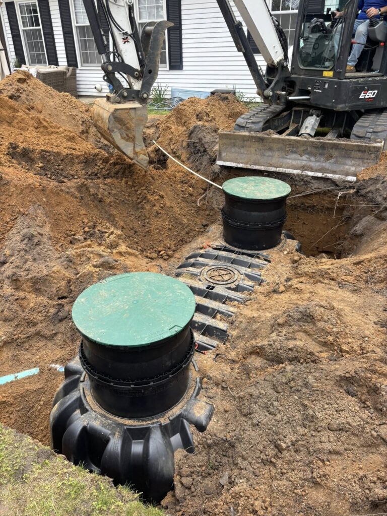 Compact excavator at a residential yard, digging a trench for a septic system beside green access lids on black tanks.