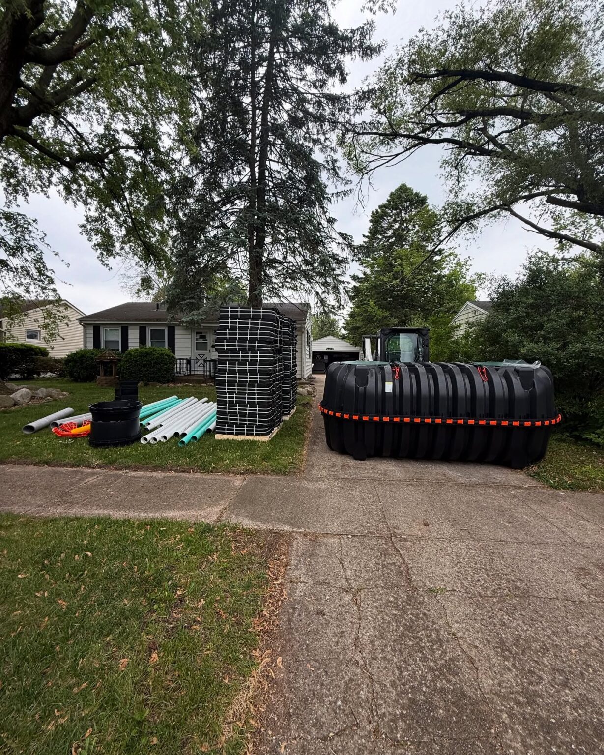 Residential front yard with stacked black septic tanks, piles of pvc pipes, and a large tree overhead, ready for installation - arwwm - arwwm