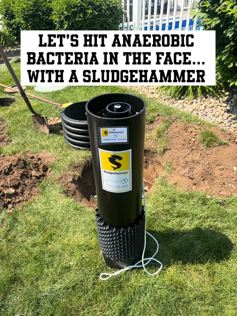 Black SludgeHammer treatment device installed upright in a dug hole in a grassy yard, with a large bold promotional banner across the top reading 'LET'S HIT ANAEROBIC BACTERIA IN THE FACE... WITH A SLUDGEHAMMER' and a shovel nearby nearby.