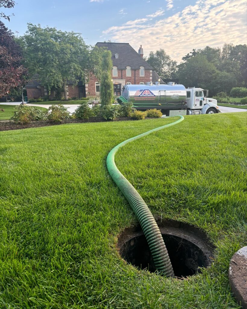 Green corrugated sewer hose extends from an open manhole across a well-manicured lawn toward a white service truck and brick house in a suburban yard. - arwwm - arwwm