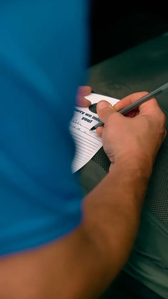 Documentation - ARWWM - ARWWM Person writes on a small card that says 'sorry we miss you' with a pen, close-up on hands and blue clothing. - arwwm - arwwm