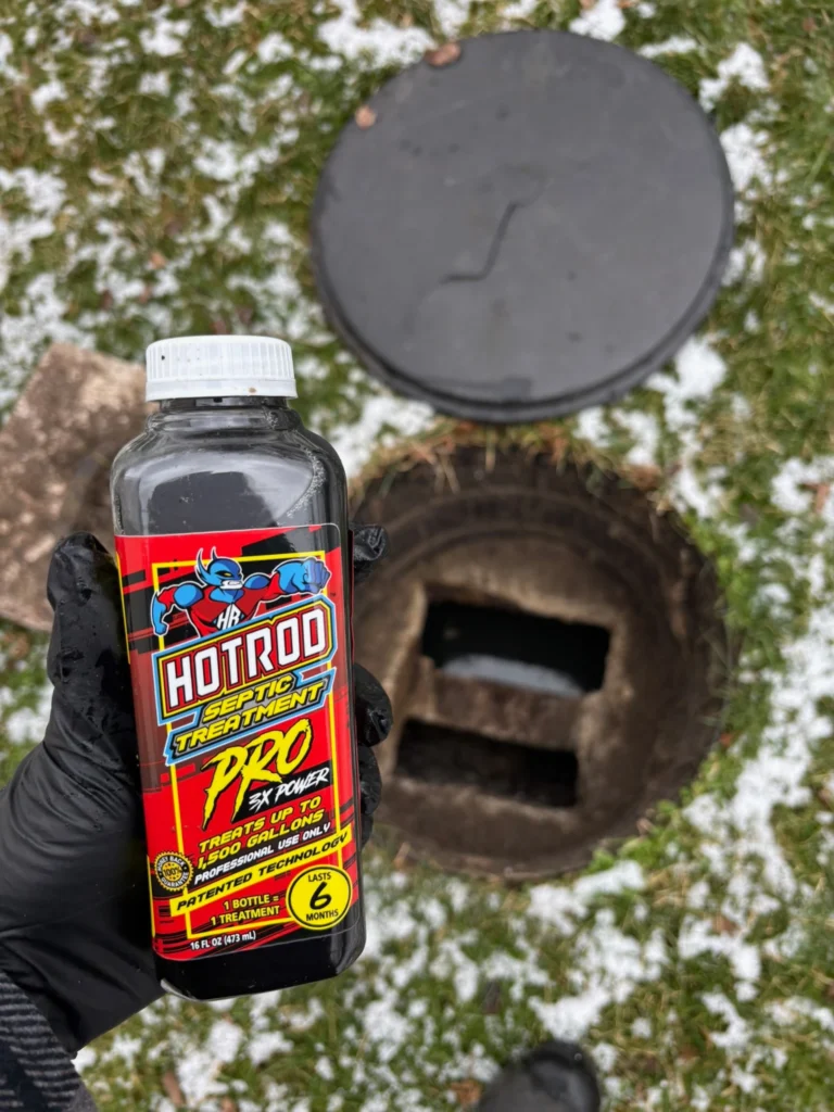 Gloved hand holding a red hotrod septic treatment bottle beside a dark round septic tank lid outdoors on a snowy, grassy surface. ", - arwwm - arwwm