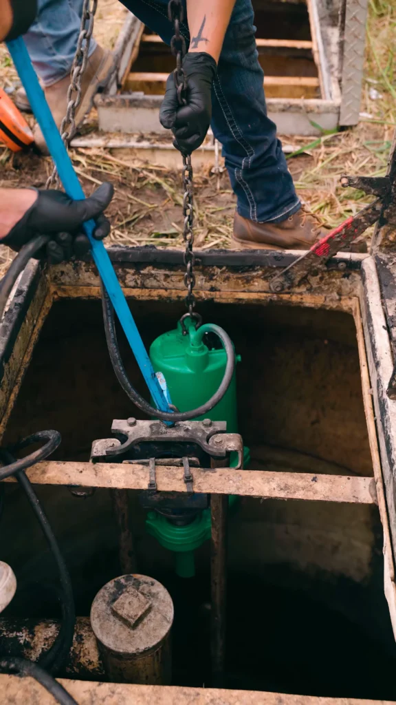 Worker in gloves lowering a green water pump into a dirty open well using a chain and blue pipe. - arwwm - arwwm