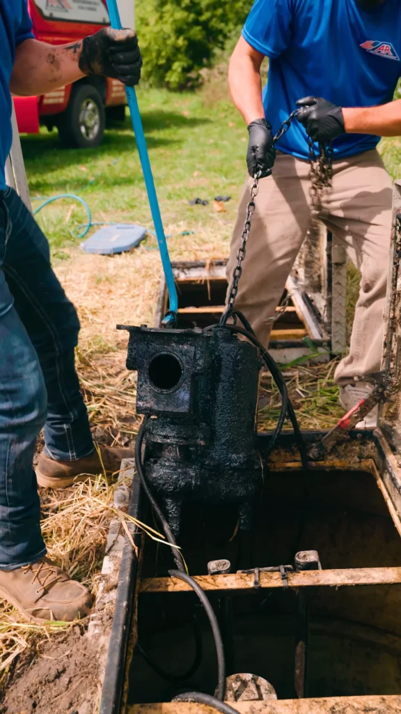 Two workers in gloves stand by an open manhole, lowering a chained black pump or apparatus into a dark underground shaft. - arwwm - arwwm