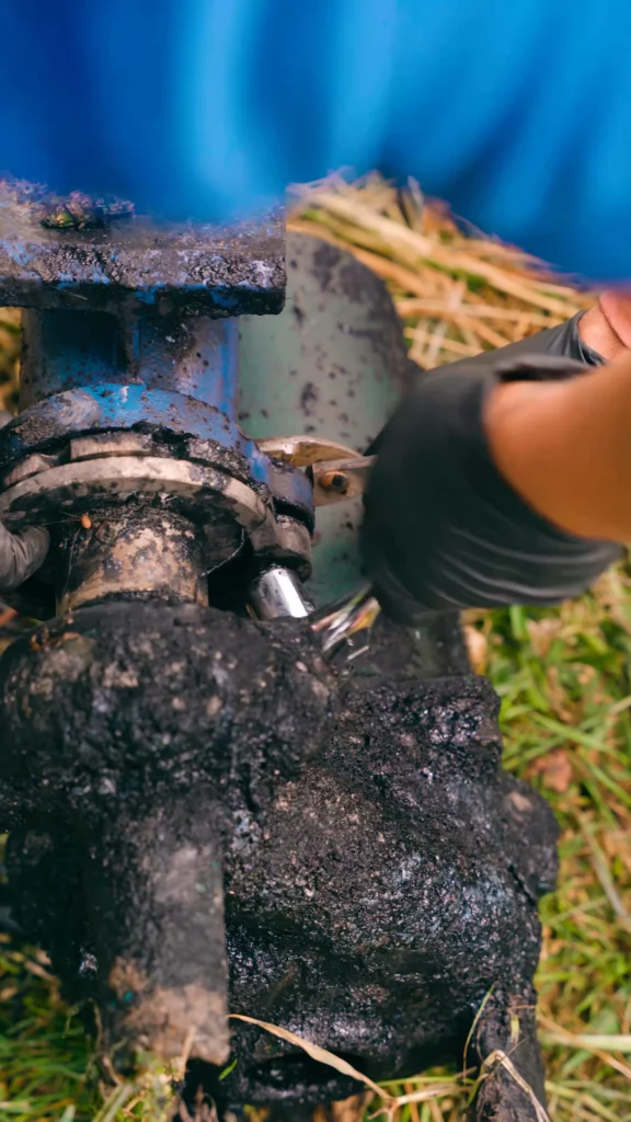 Pump Repair 2 - ARWWM - ARWWM Close-up of a corroded valve on a blue-painted pipe, covered in black grease, with a gloved hand nearby outdoors. - arwwm - arwwm