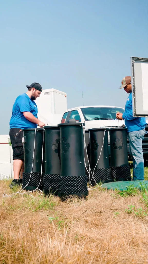 Sludgehammer Install 1 - ARWWM - ARWWM Two workers in blue shirts stand beside a row of black recycling bins connected with white cables at an outdoor waste site, with a white truck in the background. - arwwm - arwwm