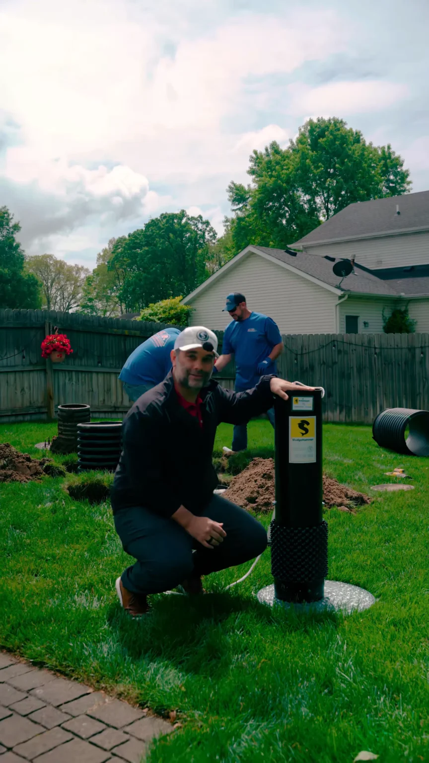 Sludgehammer explained - ARWWM - ARWWM Man kneels in a backyard beside a dark drainage utility post during a yard drainage install, with two workers and a house in the background. - arwwm - arwwm