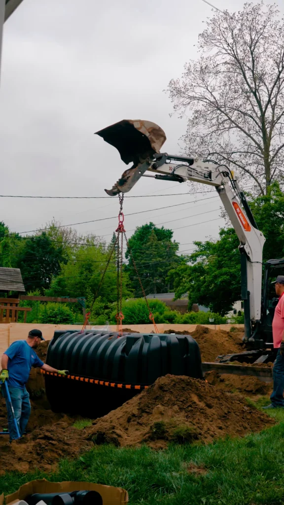 Construction site with an excavator lifting a large black corrugated pipe into a trench; workers stand nearby on dirt and grass. - arwwm - arwwm