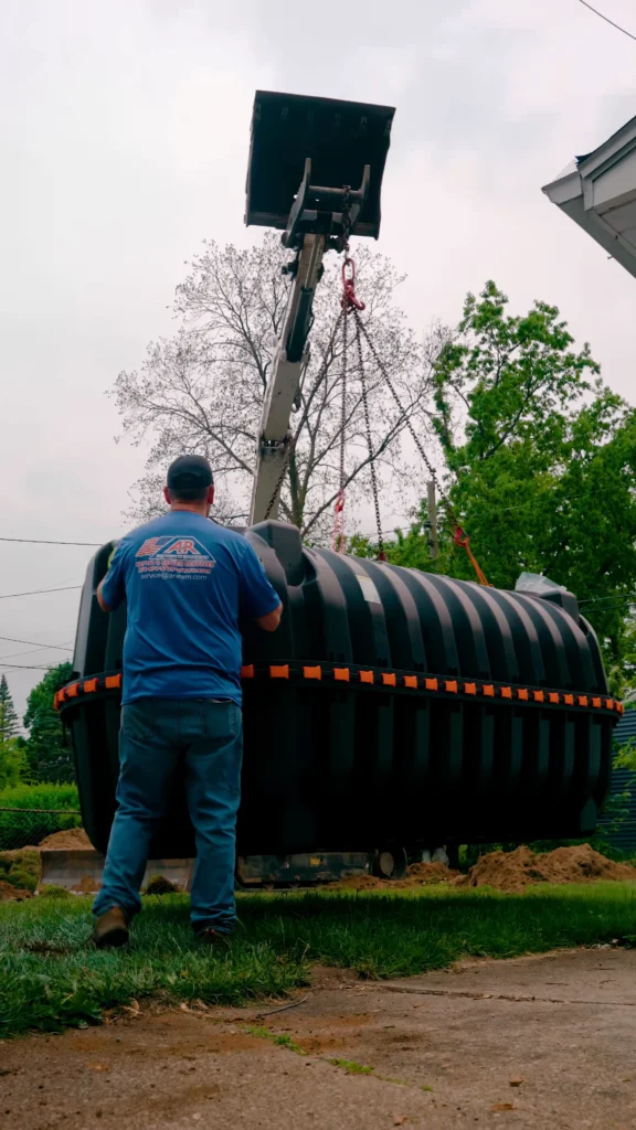 Worker in a blue shirt and cap supervises the installation of a large black ribbed underground tank as a crane lowers it into place. - arwwm - arwwm