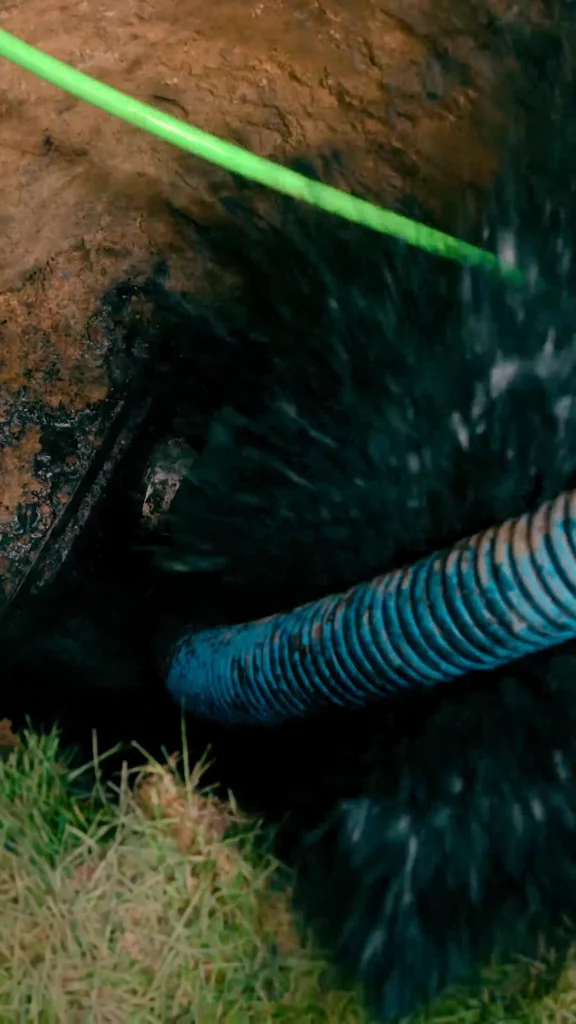 Green pipe spans a muddy excavation, with a blue corrugated hose nearby and grass at the edge. - arwwm - arwwm